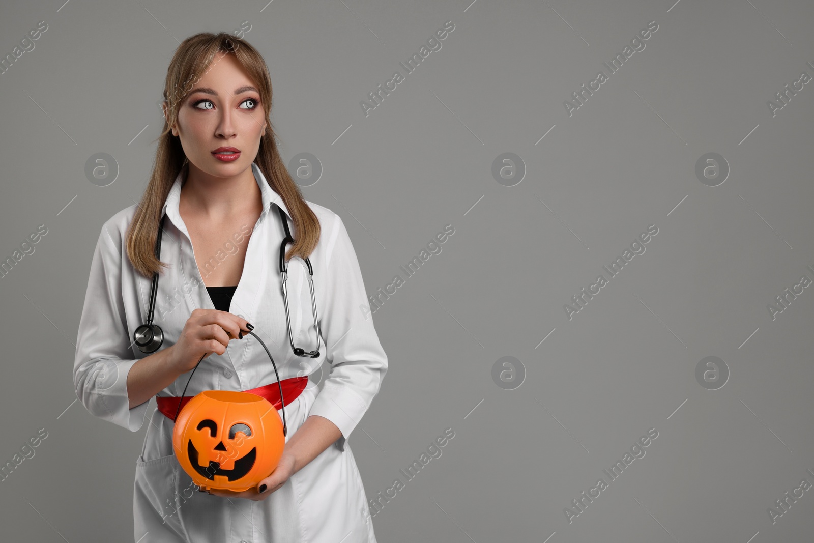 Photo of Woman in scary nurse costume with pumpkin bucket on light grey background, space for text. Halloween celebration