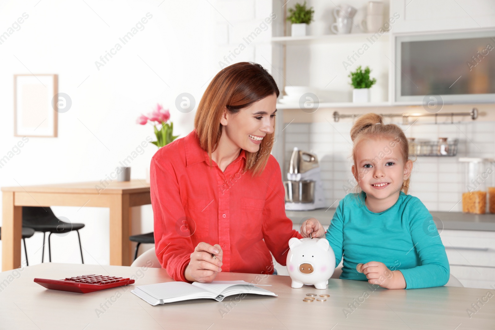 Photo of Mother and daughter putting coin into piggy bank at table indoors. Saving money