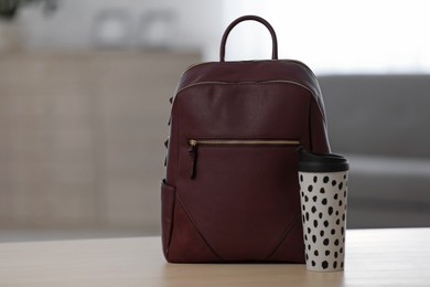 Photo of Stylish burgundy backpack and cup of coffee on wooden table indoors