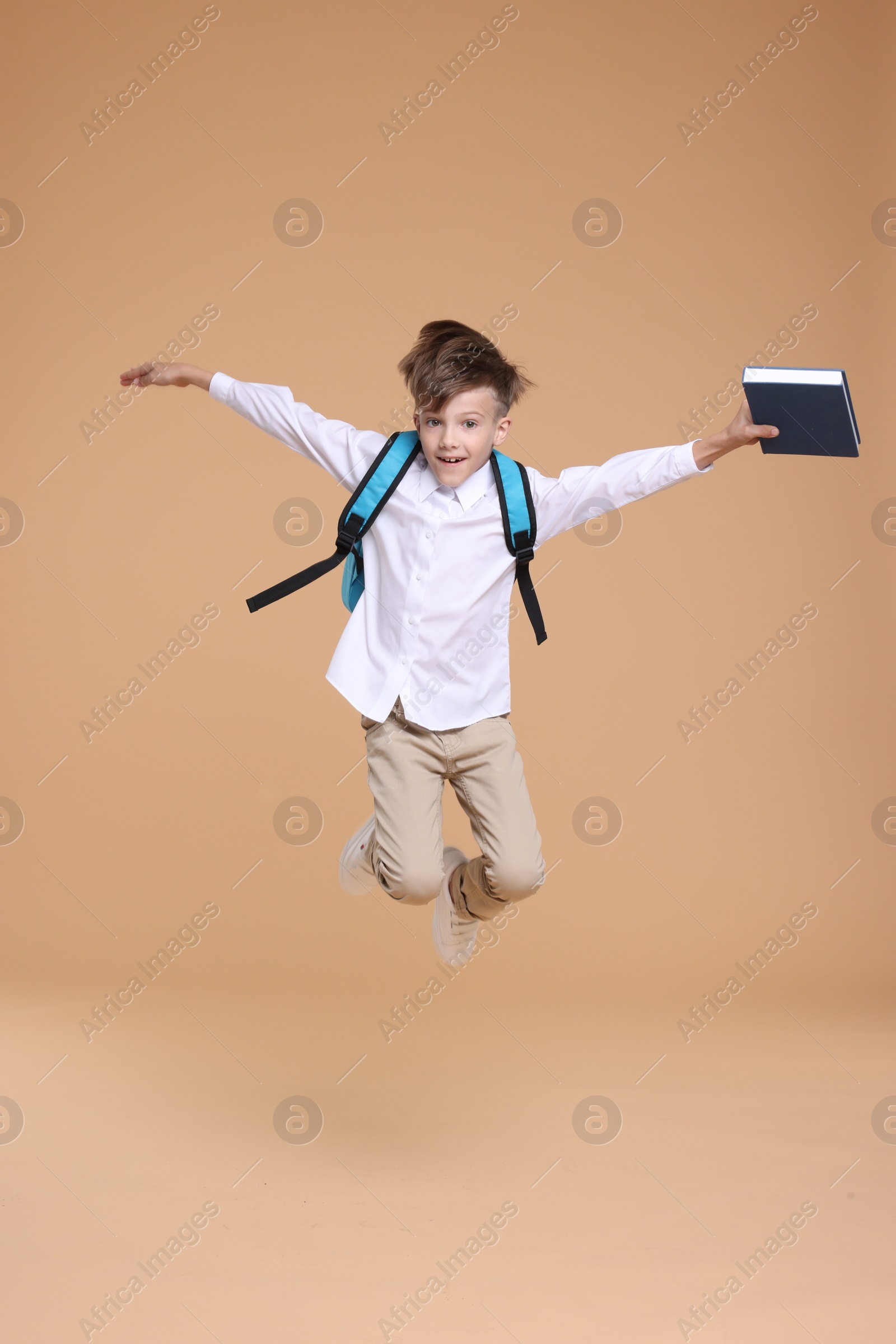 Photo of Cute schoolboy holding book and jumping on beige background, space for text