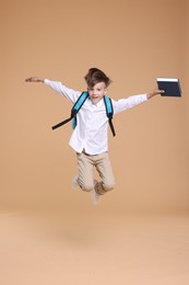 Cute schoolboy holding book and jumping on beige background, space for text