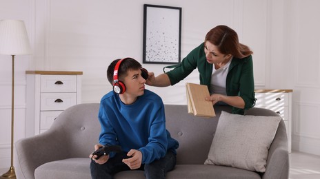 Photo of Strict mother with books scolding her son while he playing videogame at home. Teenager problems