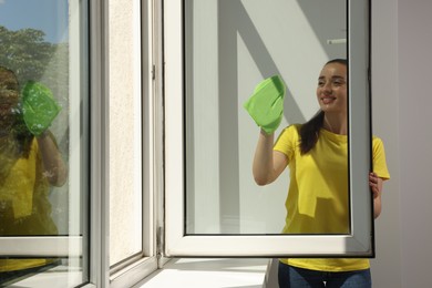 Photo of Happy young woman cleaning window glass with rag indoors