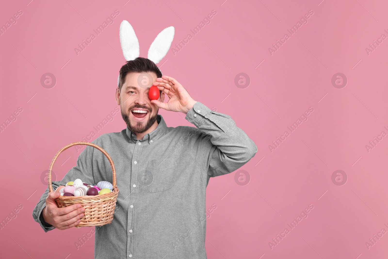 Photo of Happy man in bunny ears headband holding wicker basket with painted Easter eggs on pink background. Space for text