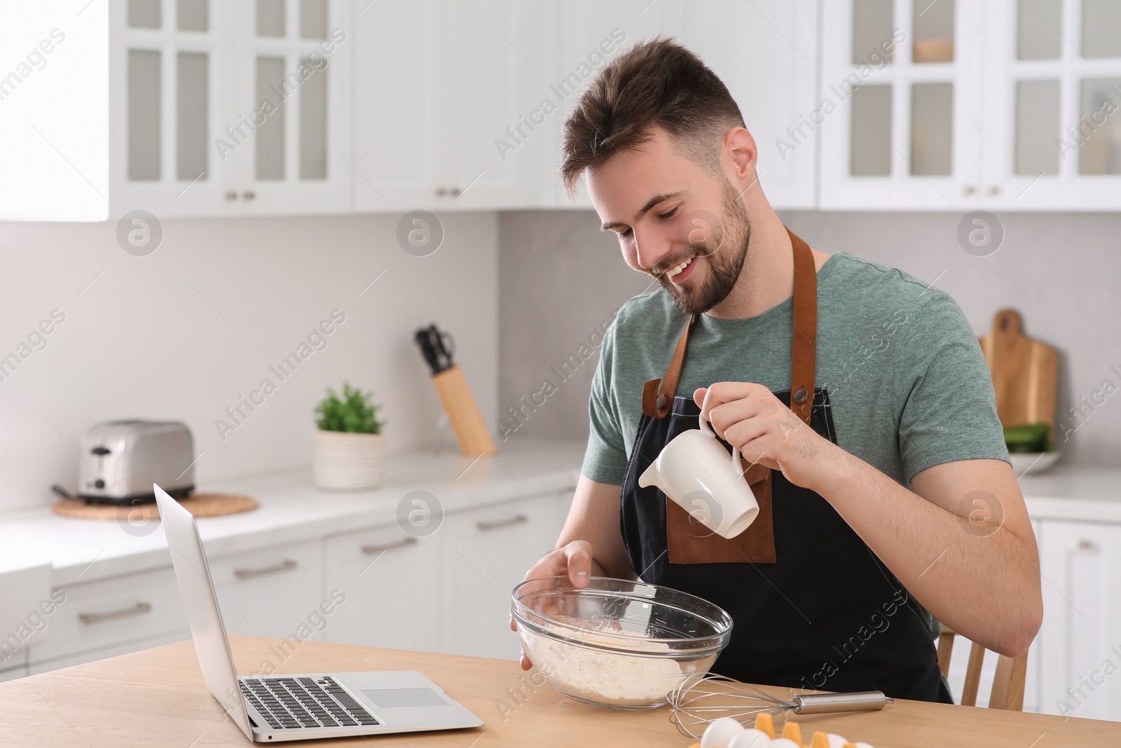 Photo of Happy man learning to cook with online video on laptop at table in kitchen. Time for hobby