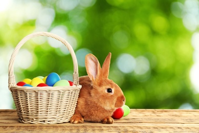 Adorable bunny and wicker basket with Easter eggs on wooden surface outdoors, space for text 