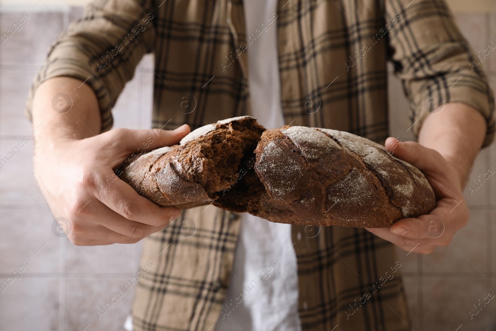 Photo of Man breaking loaf of fresh bread near grey wall, closeup