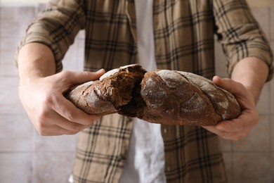 Photo of Man breaking loaf of fresh bread near grey wall, closeup