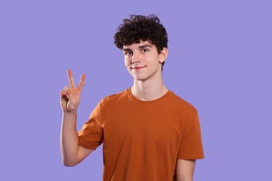 Portrait of cute teenage boy showing peace gesture on violet background