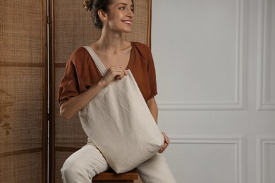 Photo of Happy young woman with blank eco friendly bag sitting on wooden stool indoors. Space for text