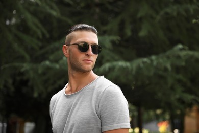 Handsome young man in stylish sunglasses at park, space for text