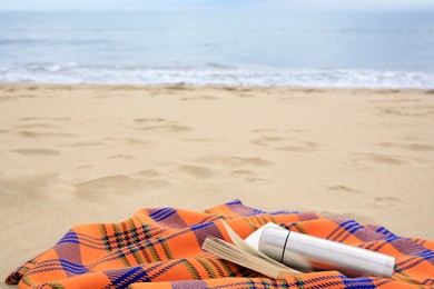 Metallic thermos with hot drink, open book and plaid on sandy beach near sea, space for text