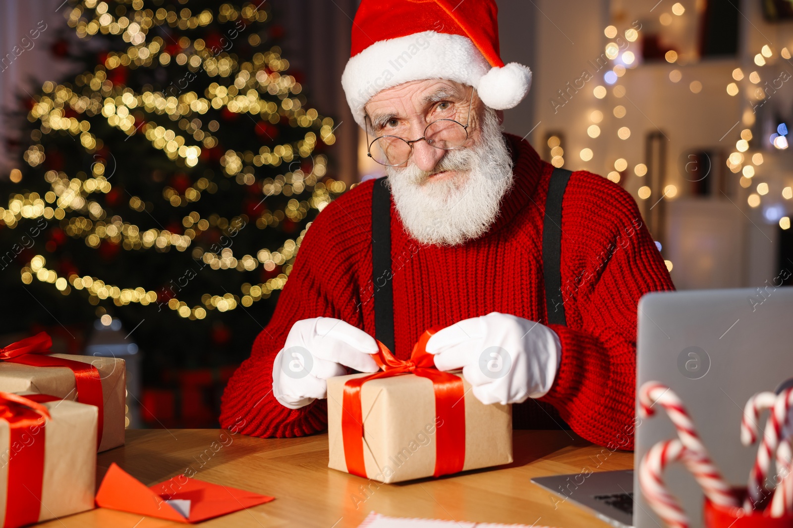 Photo of Santa Claus tying bow on gift box at his workplace in room decorated for Christmas