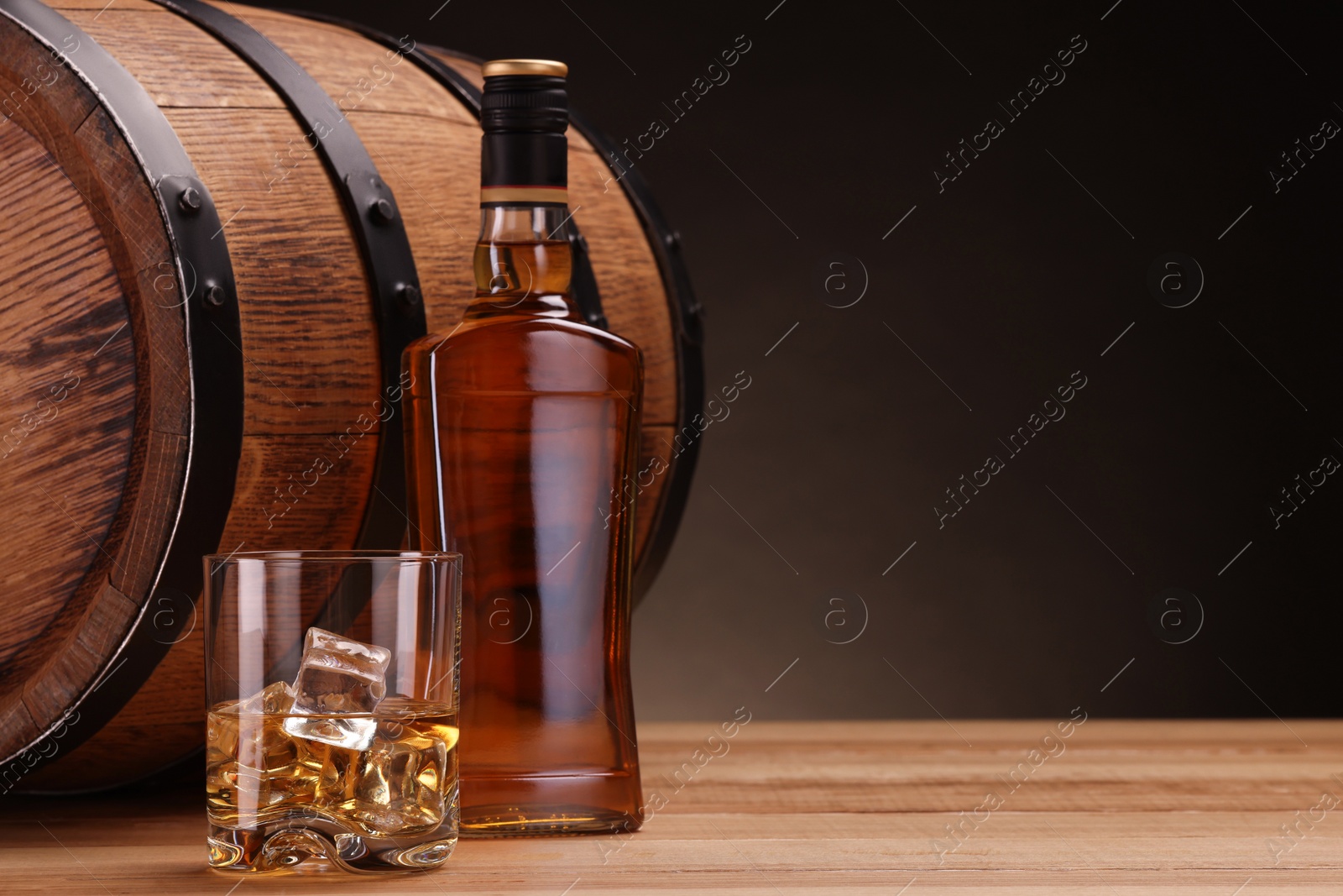 Photo of Whiskey with ice cubes in glass, bottle and barrel on wooden table against dark background, space for text