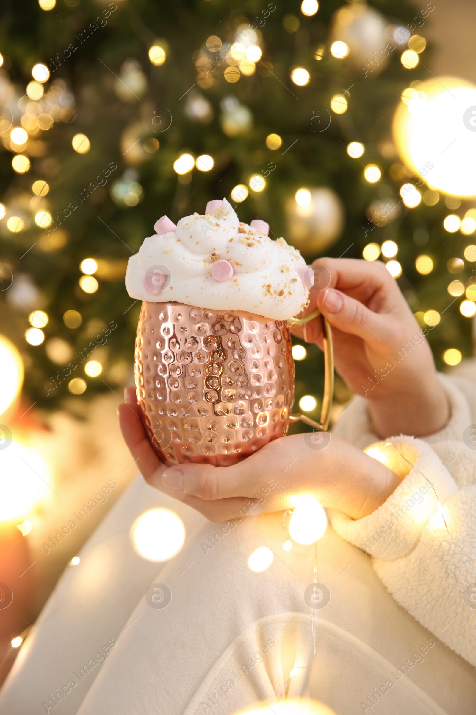 Photo of Woman holding cup of delicious drink with whipped cream and marshmallows near Christmas tree indoors, closeup