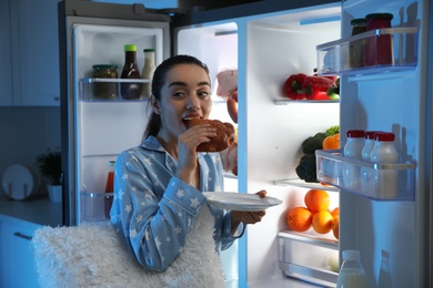 Photo of Young woman eating bun near open refrigerator at night
