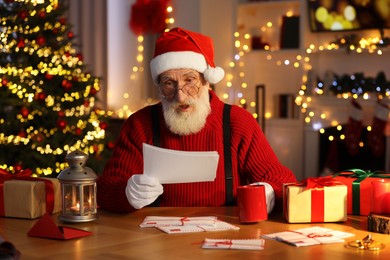 Photo of Santa Claus reading letter and drinking hot beverage at his workplace in room decorated for Christmas