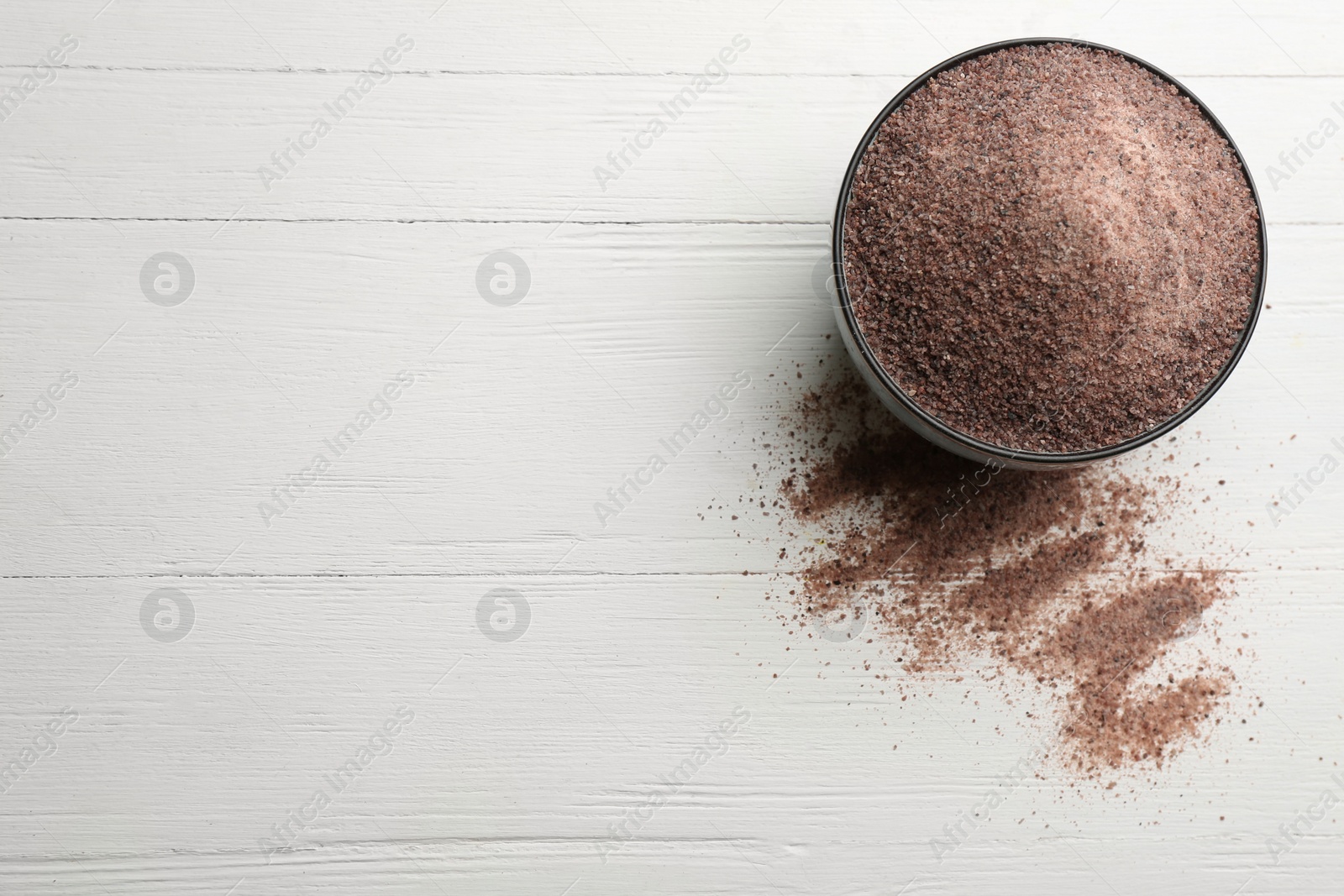 Photo of Ground black salt in bowl on white wooden table, top view. Space for text