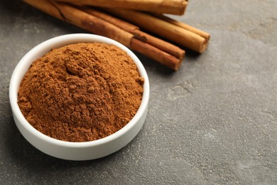 Photo of Bowl of cinnamon powder and sticks on grey table, closeup. Space for text