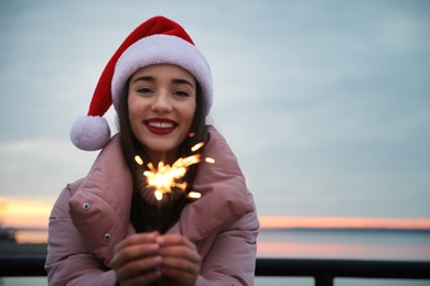 Photo of Woman in Santa hat holding burning sparkler near river, space for text