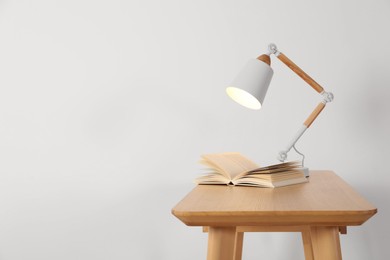 Stylish modern desk lamp and open book on wooden table near white wall, space for text