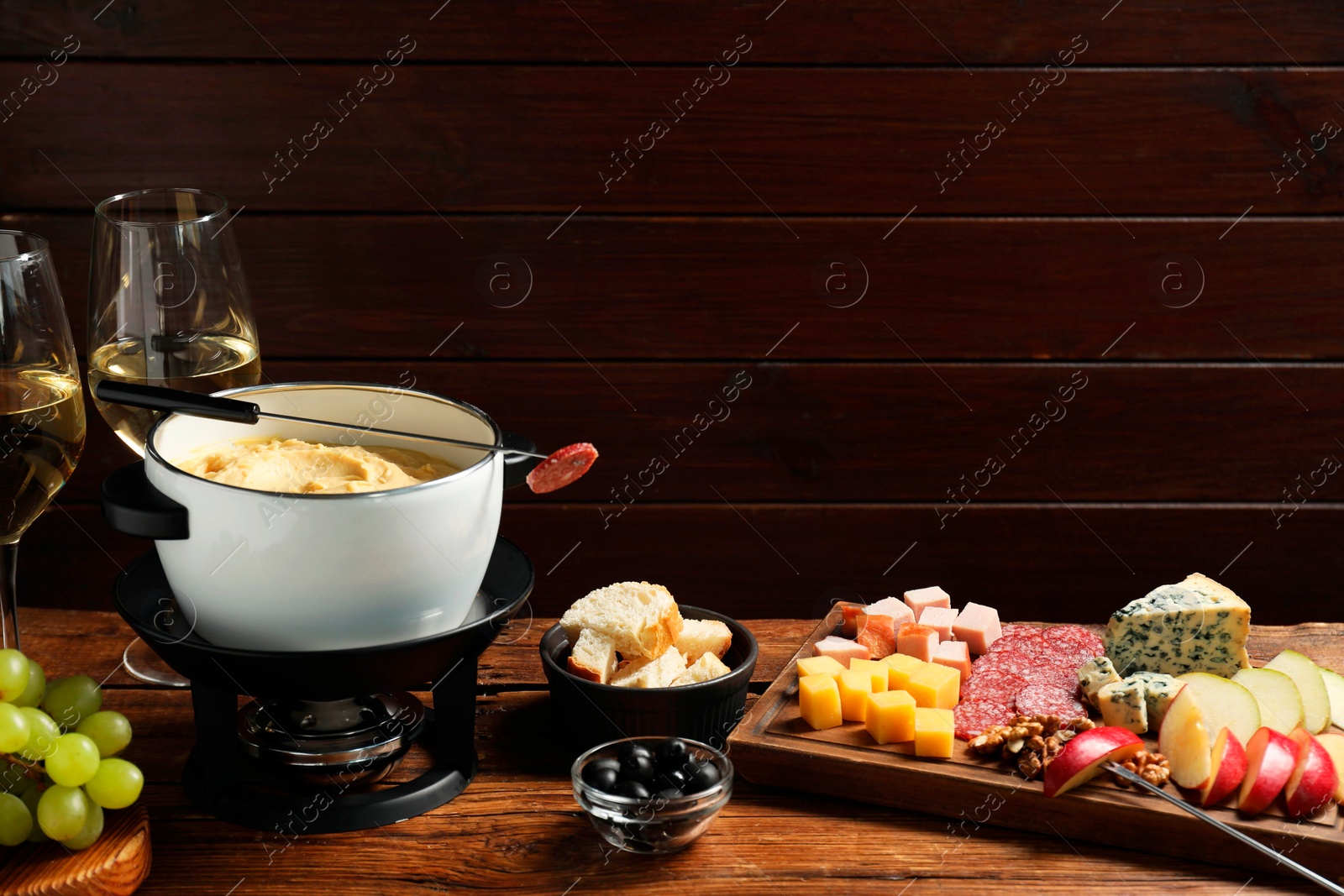 Photo of Fork with piece of sausage, melted cheese in fondue pot and other products on wooden table. Space for text