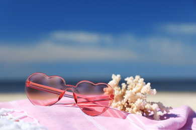 Photo of Stylish heart shaped sunglasses, coral and blanket on sand near sea, closeup. Beach accessories for summer vacation