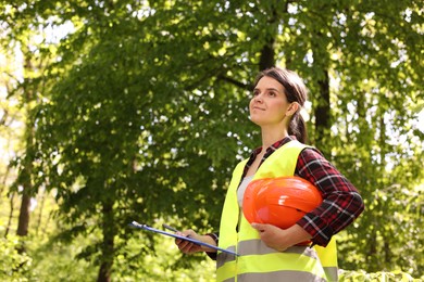 Forester with hard hat and clipboard examining plants in forest, space for text