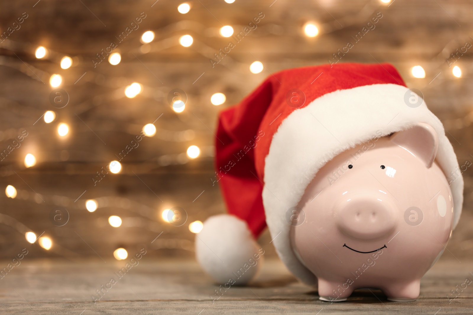 Photo of Piggy bank with Santa hat on table against blurred Christmas lights. Space for text