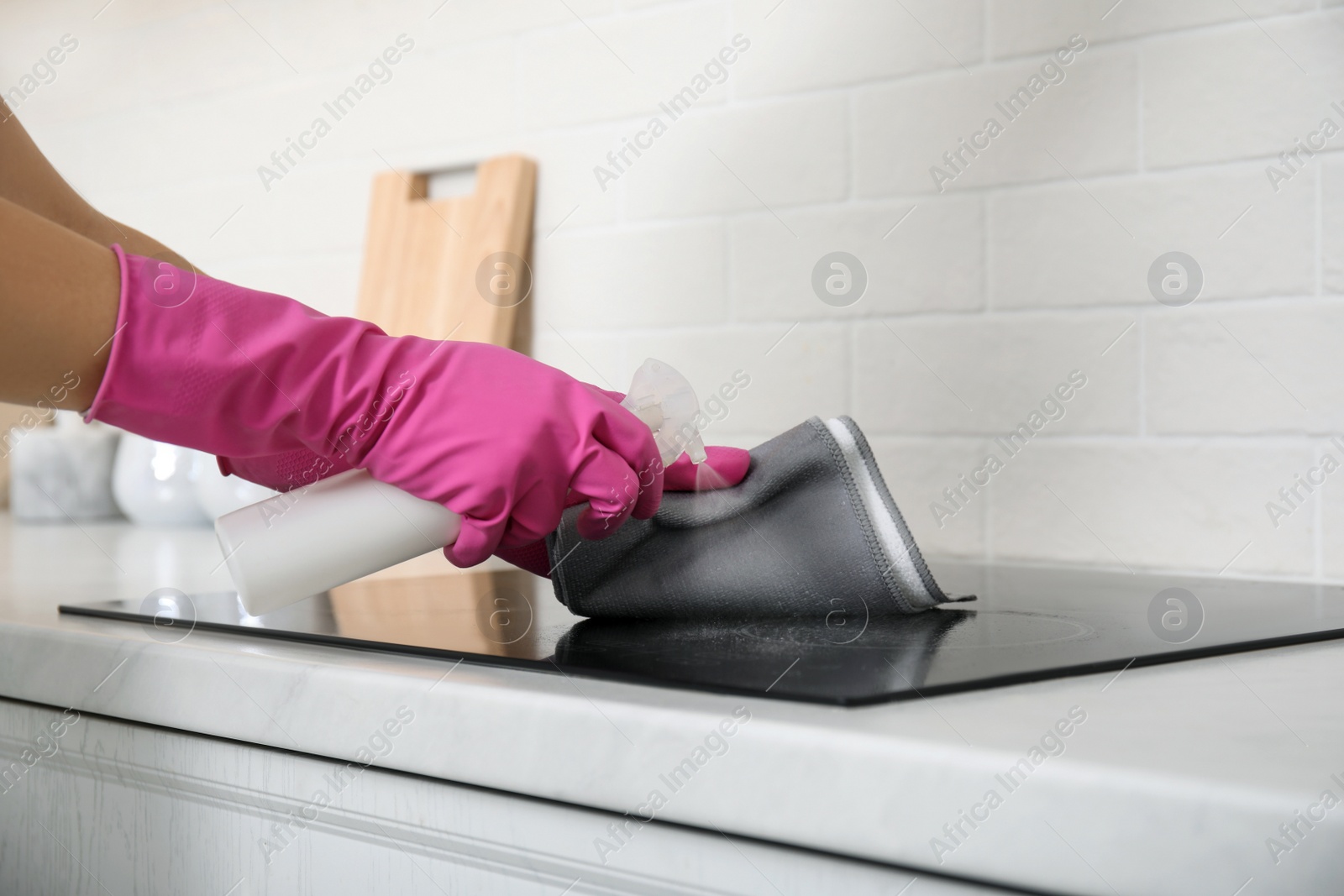 Photo of Woman cleaning stove with rag and detergent in kitchen, closeup