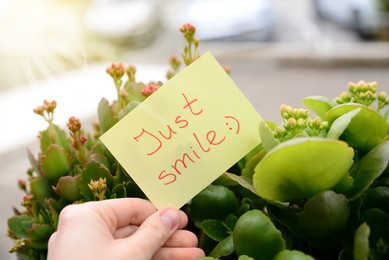 Photo of Woman holding paper note with handwritten text Just smile near beautiful plants against blurred background, closeup