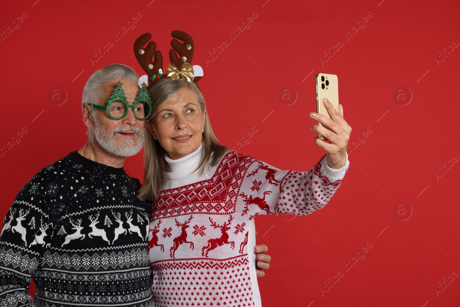 Photo of Senior couple in Christmas sweaters, reindeer headband and party glasses taking selfie on red background. Space for text