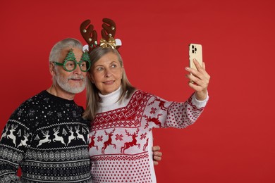 Photo of Senior couple in Christmas sweaters, reindeer headband and party glasses taking selfie on red background. Space for text