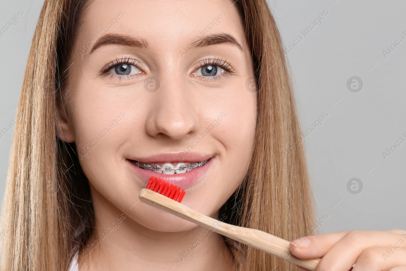 Photo of Smiling woman with dental braces cleaning teeth on grey background, closeup