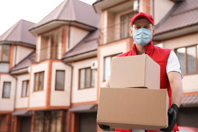 Photo of Courier in protective mask and gloves with boxes near house outdoors. Delivery service during coronavirus quarantine