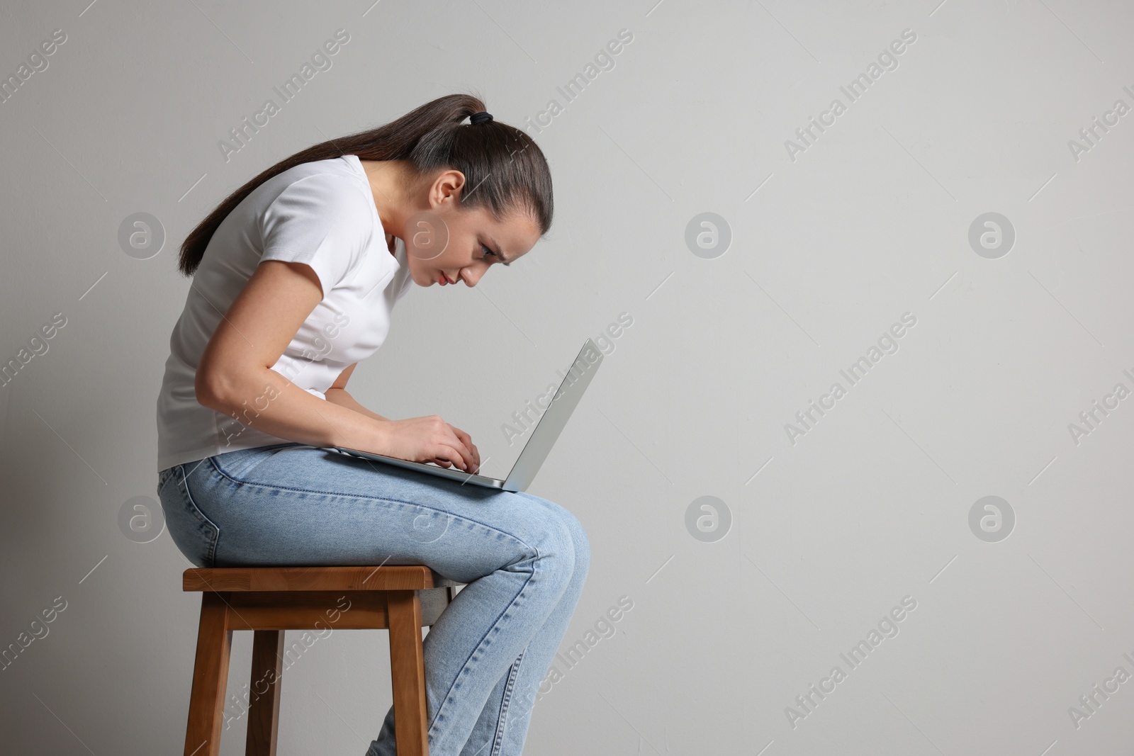 Photo of Young woman with poor posture using laptop while sitting on stool against grey background, space for text