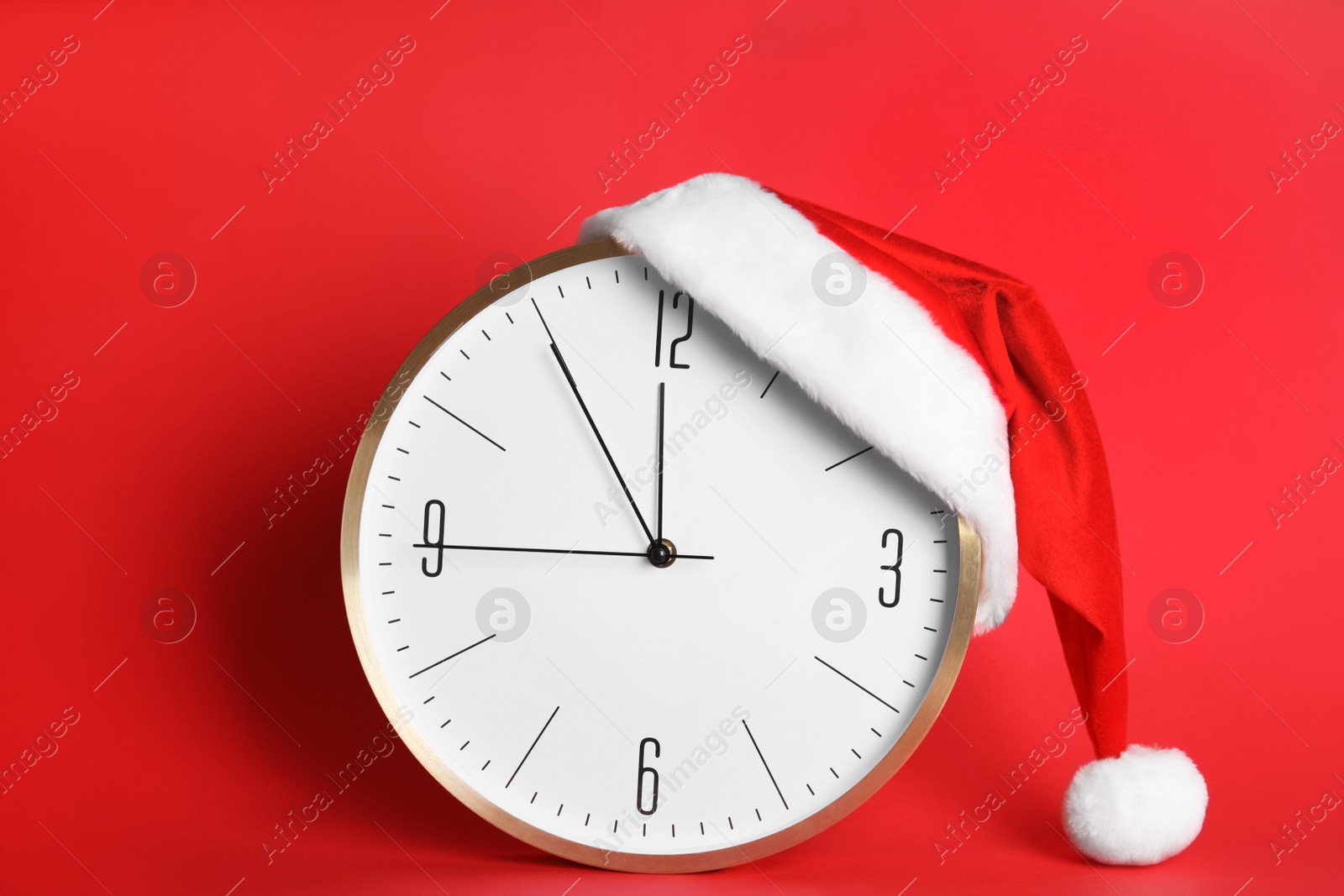 Photo of Clock with Santa hat showing five minutes until midnight on red background. New Year countdown