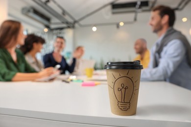 Photo of Team of employees working together in office. Paper cup of drink on white table, selective focus