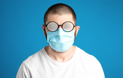 Photo of Man with foggy glasses caused by wearing disposable mask on blue background. Protective measure during coronavirus pandemic