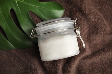 Jar of salt scrub and palm leaf on brown towel, flat lay