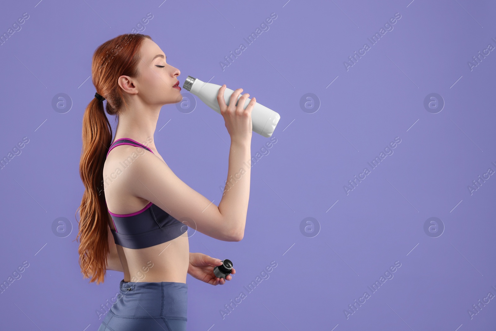Photo of Woman in sportswear drinking from thermo bottle on violet background, space for text