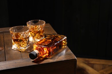 Photo of Whiskey with ice cubes in glasses and bottle on wooden crate against black background, closeup. Space for text