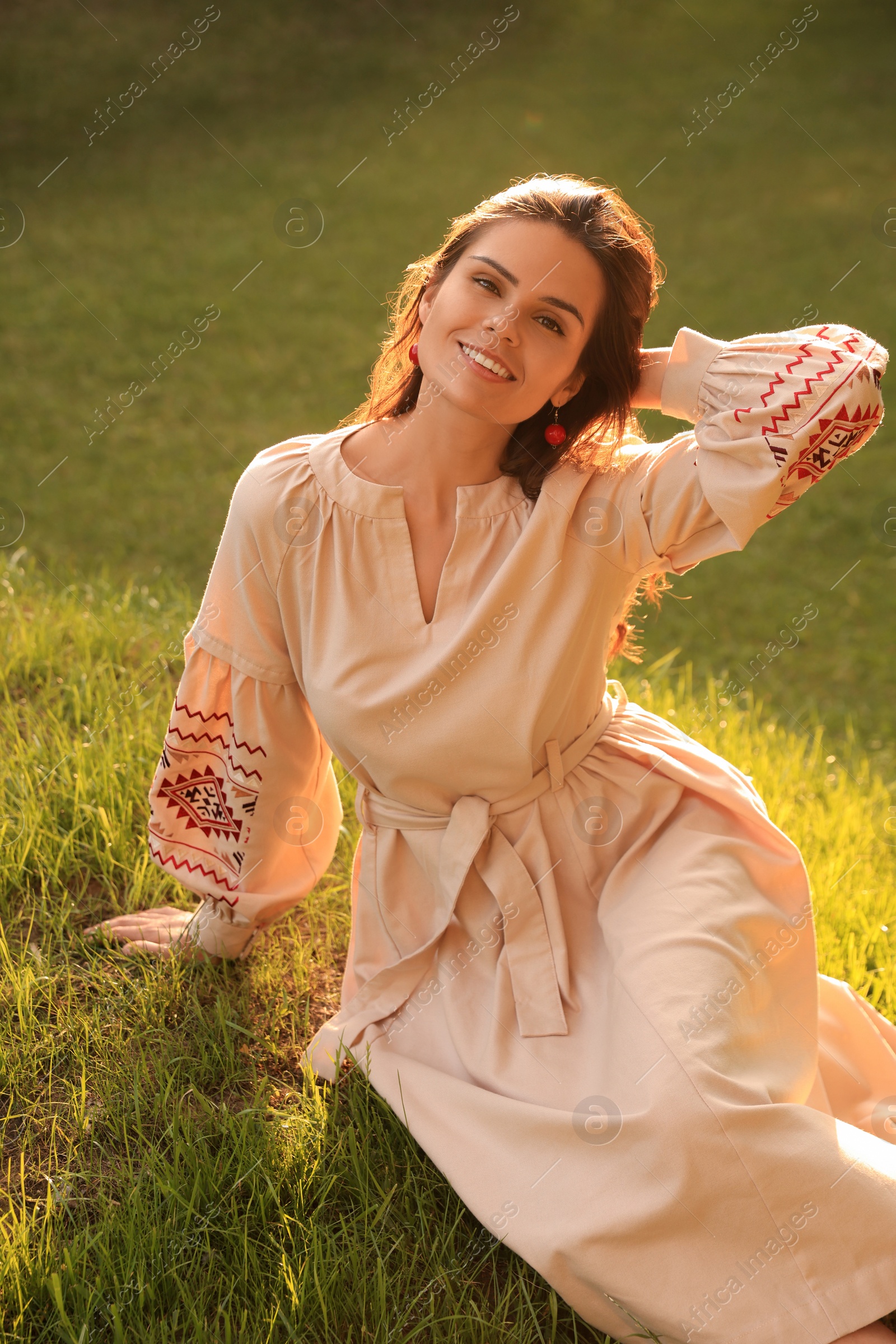 Photo of Beautiful woman in embroidered dress sitting on green grass outdoors. Ukrainian national clothes