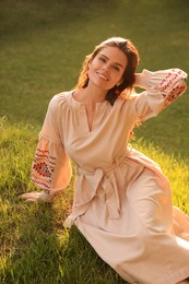 Photo of Beautiful woman in embroidered dress sitting on green grass outdoors. Ukrainian national clothes