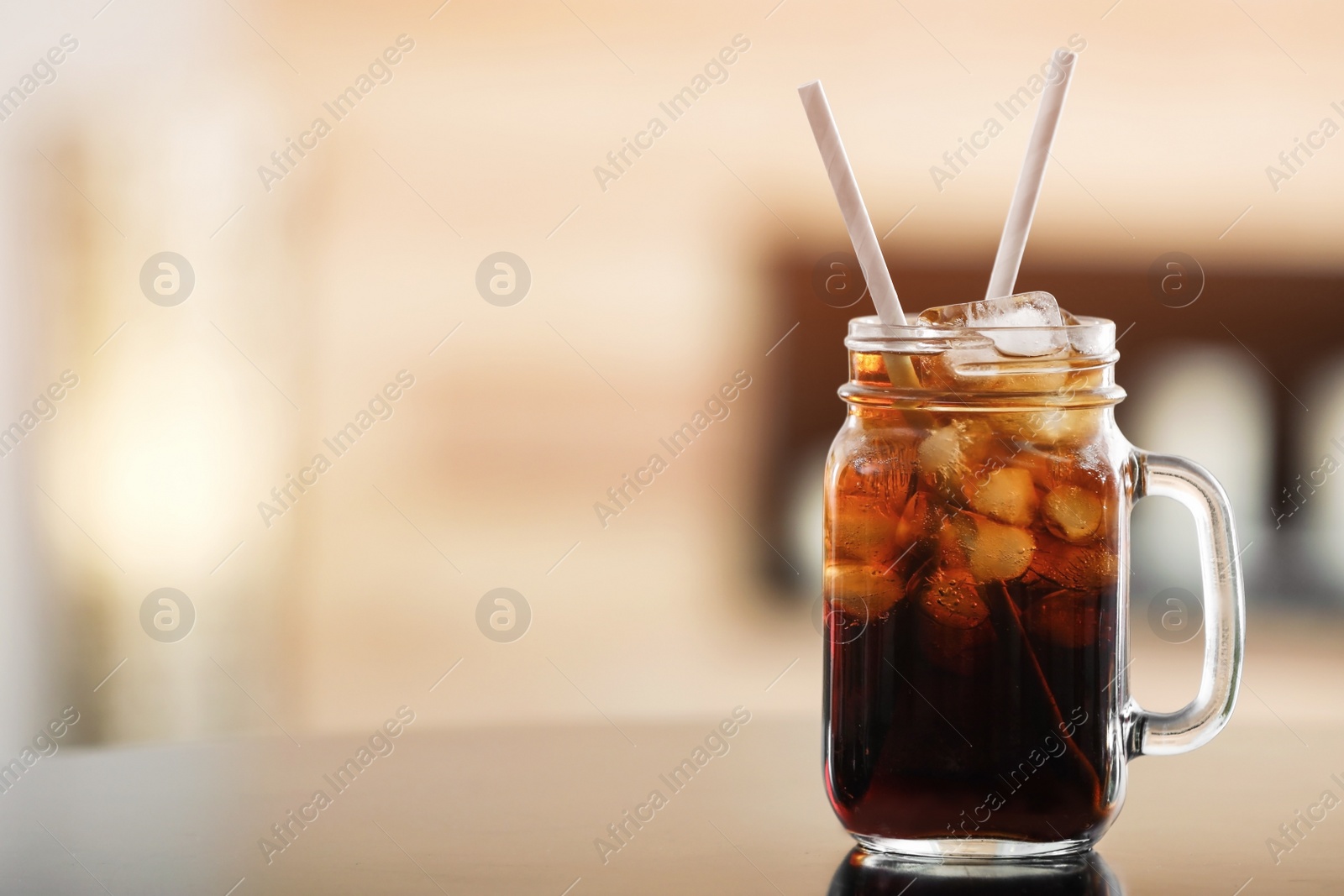 Photo of Mason jar of cola with ice on table against blurred background. Space for text
