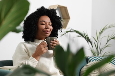 Photo of Relaxing atmosphere. Woman with cup of hot drink near beautiful houseplants indoors
