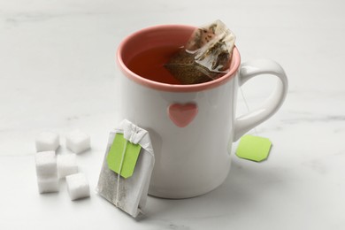 Photo of Tea bags, cup of hot beverage and sugar cubes on white table, closeup