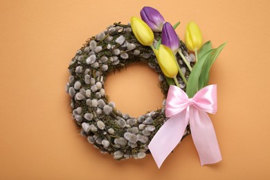 Photo of Wreath made of beautiful willow, colorful tulip flowers and pink bow on orange background, top view