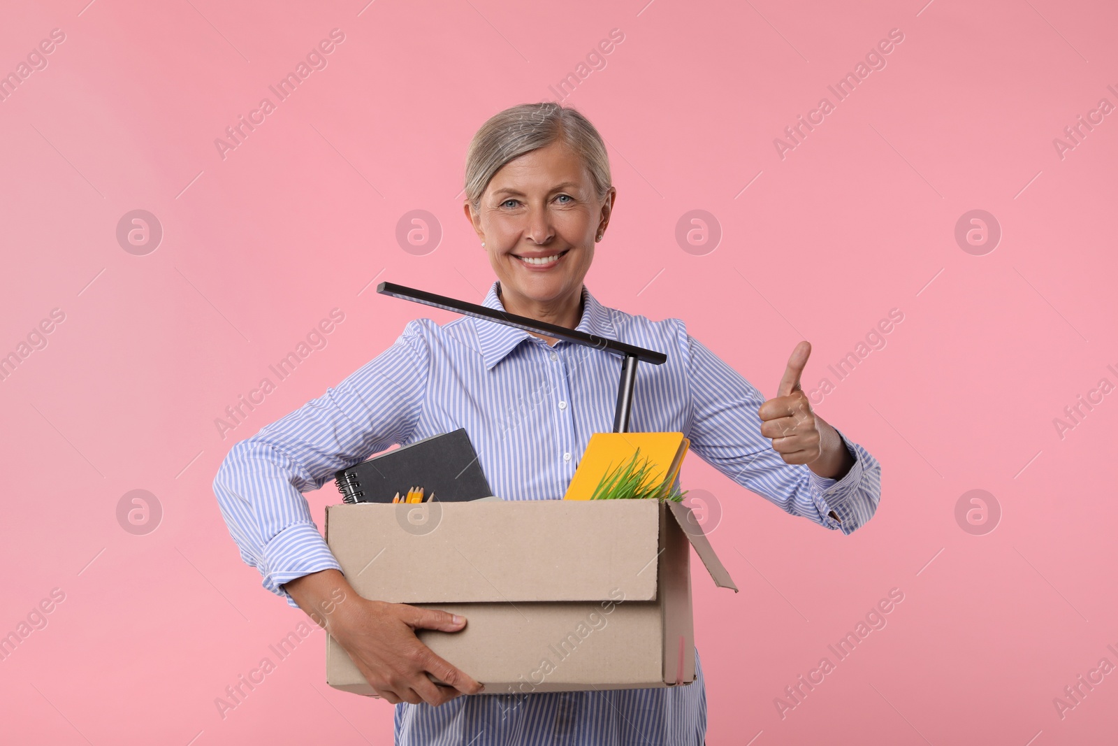 Photo of Happy unemployed senior woman with box of personal office belongings showing thumb up on pink background