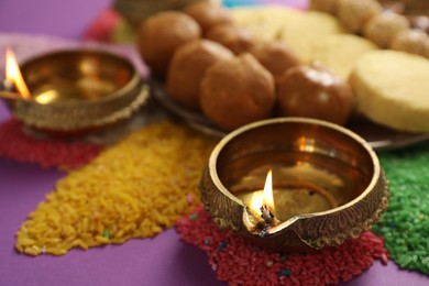 Diwali celebration. Tasty Indian sweets, diya lamps and colorful rangoli on violet table, closeup